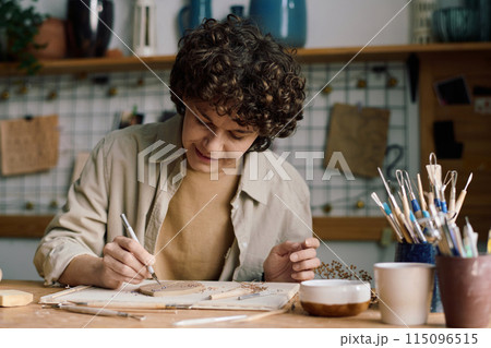 Creative Caucasian woman sitting at table in pottery workshop creating leaf-shaped plate, copy space 115096515