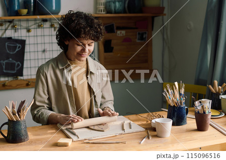Medium shot of mature Caucasian woman sitting at table in workshop pressing dried flowers to clay leaf using rolling pin 115096516
