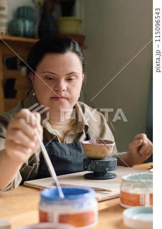 Young woman with Down syndrome sitting at table taking blue paint with paintbrush to color handmade vessel 115096543