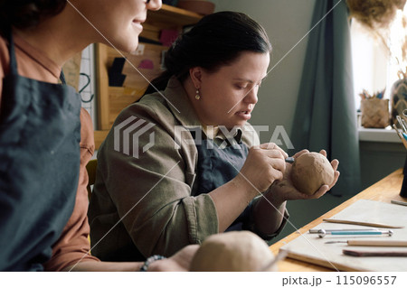 Selective focus shot of young woman with Down syndrome using clay carving tools and chatting with her friend during pottery class Selective focus shot of young woman with Down syndrome using clay carving tools and chatting with her friend during pottery class 115096557
