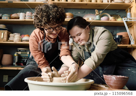 Low angle view shot of two diverse women wearing aprons having fun during pottery class shaping clay on wheel together Low angle view shot of two diverse women wearing aprons having fun during pottery class shaping clay on wheel together 115096568
