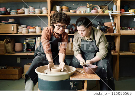 Mature Caucasian artisan demonstrating young female student with Down syndrome how to shape clay on potters wheel during class Mature Caucasian artisan demonstrating young female student with Down syndrome how to shape clay on potters wheel during class 115096569