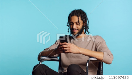 BIPOC man in wheelchair discussing with friend over online videocall using mobile phone. Person with paraplegia talking with mate in videoconference using cellphone, studio background, camera A 115097426