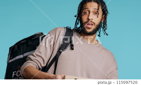 Portrait of BIPOC courier delivering pizza order to client. Delivery part time worker using thermal backpack to store food and bring it to customer, isolated over studio background, camera B Portrait of BIPOC courier delivering pizza order to client. Delivery part time worker using thermal backpack to store food and bring it to customer, isolated over studio background, camera B 115097520