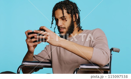 Happy man with disability in wheelchair playing videogames on cellphone, celebrating victory. Cheerful gamer enjoying game on phone, excited about win, studio background, camera B 115097538