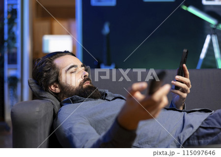 Homeowner bored at home laying down, using TV to watch movies on streaming services and chatting with friends. Apathetic man lounged on couch, mindlessly consuming entertainment and using mobile phone Homeowner bored at home laying down, using TV to watch movies on streaming services and chatting with friends. Apathetic man lounged on couch, mindlessly consuming entertainment and using mobile phone 115097646