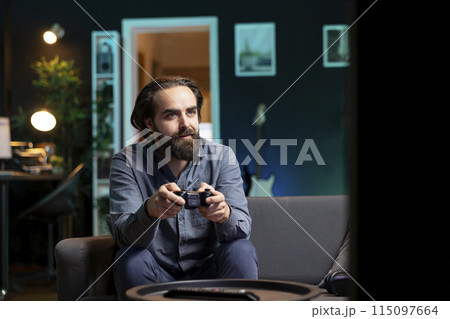 Joyous man playing videogame in home theatre, holding controller. Smiling gamer participating in PvP online multiplayer game using console system and high quality gamepad Joyous man playing videogame in home theatre, holding controller. Smiling gamer participating in PvP online multiplayer game using console system and high quality gamepad 115097664