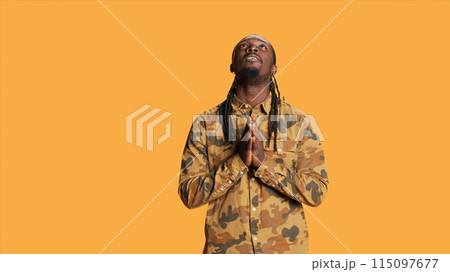 African american person praying to Jesus in studio, being spiritual and showing belief in God over orange background. Young adult saying prayer and feeling hopeful, religious man smiling. 115097677