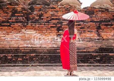 Young woman wearing a red Thai dress and gold accessories stands holding an ancient umbrella with her back turned in the ancient area of Wat Chaiwatthanaram, Ayutthaya, Thai national costume. 115099490