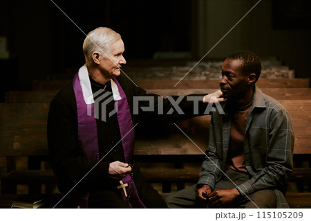 Medium shot of senior Caucasian Catholic priest and young African American man sitting on pew in church having conversation Medium shot of senior Caucasian Catholic priest and young African American man sitting on pew in church having conversation 115105209