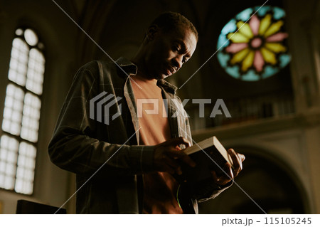 Low angle view medium shot of African American man standing in Catholic church reading religious text in book Low angle view medium shot of African American man standing in Catholic church reading religious text in book 115105245