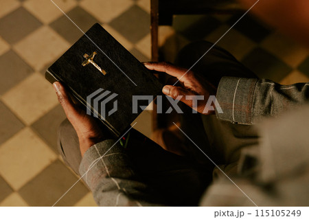 High angle view of hands of unrecognizable Black man sitting on pew in Catholic church holding prayer book High angle view of hands of unrecognizable Black man sitting on pew in Catholic church holding prayer book 115105249