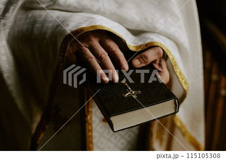 Closeup of wrinkled hands of unrecognizable senior Catholic priest holding Holy Bible book 115105308