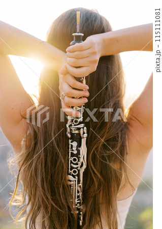Girl with a clarinet in her hands behind her head stands in the sun. Back view 115111081