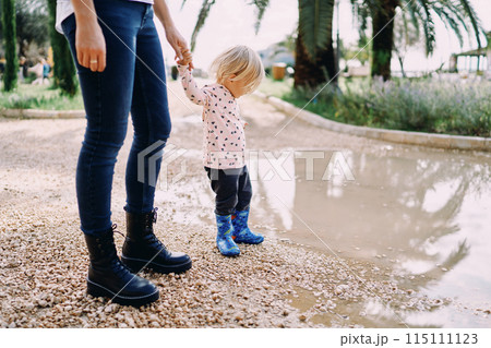 Mom holds the hand of a little girl in rubber boots standing on the edge of a large puddle Mom holds the hand of a little girl in rubber boots standing on the edge of a large puddle 115111123