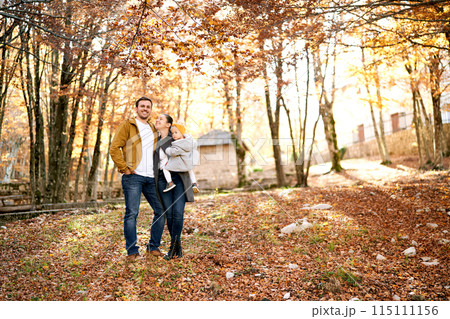 Smiling mom with little girl in her arms looks at dad hugging her in the park Smiling mom with little girl in her arms looks at dad hugging her in the park 115111156