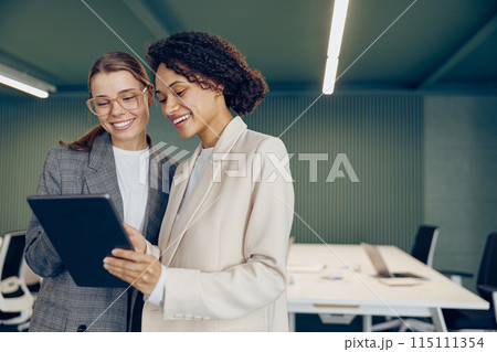 Two busy female coworkers discussing project standing in modern office and use digital tablet Two busy female coworkers discussing project standing in modern office and use digital tablet 115111354