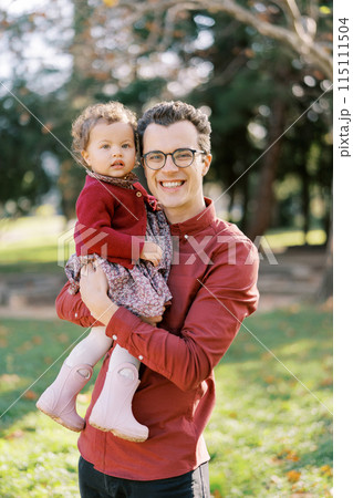 Smiling dad with a little girl in his arms stands in the park 115111504