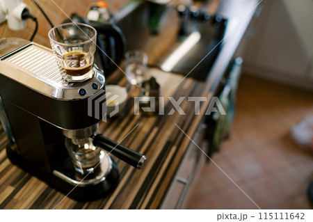 Glass of coffee stands on a carob coffee machine preparing coffee in a cup Glass of coffee stands on a carob coffee machine preparing coffee in a cup 115111642