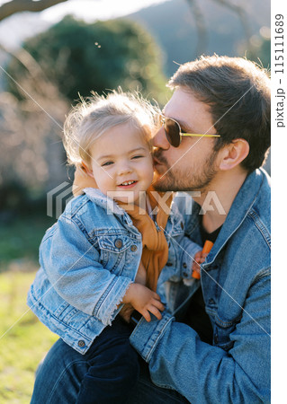 Dad kisses on the cheek a little smiling girl on his knees sitting on the lawn 115111689