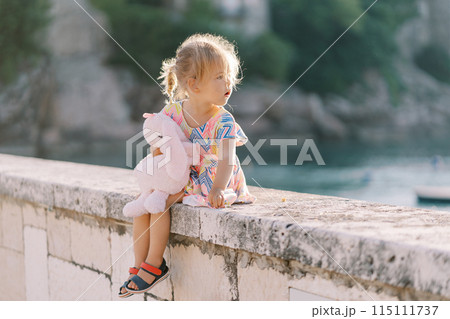 Little girl sits hugging a plush hare on a stone fence, looking back 115111737