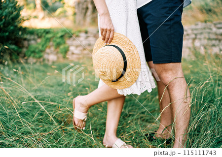 Straw hat in the hand of woman standing next to man in the garden. Cropped. Faceless 115111743