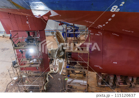 Cargo vessel in dry dock on ship repairing yard. Variable pitch propeller and rudder. Welder working on ship repairing yard. 115120637