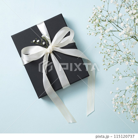 a black gift box with a white ribbon on a blue background with a bouquet of  gypsophila.  115120737