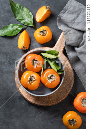Fresh ripe persimmon fruits in bowl on a cutting board on a dark background with slices and leaves. Fresh ripe persimmon fruits in bowl on a cutting board on a dark background with slices and leaves. 115121150