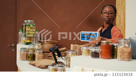Retailer weighting goods on electronic scale at checkout, serving regular client with natural groceries from local supermarket. African american worker selling organic products. Handheld shot. 115123018