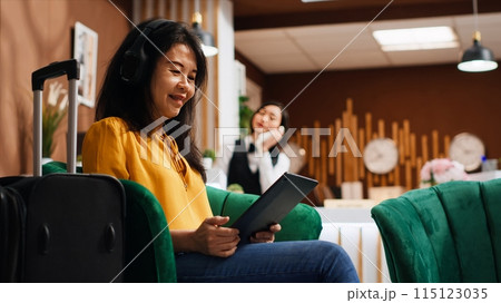Asian tourist working on tablet in lounge area at resort, relaxing and waiting to confirm room reservation and do check in. Guest scrolling online websites on device, tourism concept. Handheld shot. Asian tourist working on tablet in lounge area at resort, relaxing and waiting to confirm room reservation and do check in. Guest scrolling online websites on device, tourism concept. Handheld shot. 115123035