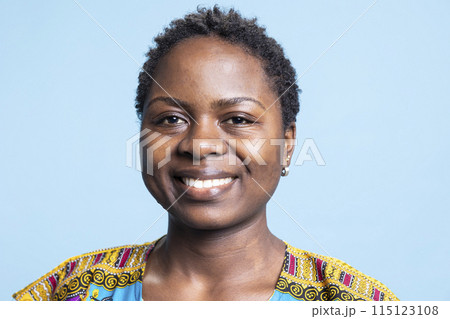 Portrait of cheerful female model posing over blue background, smiling while she wears traditional ethnic clothing. African american girl feeling confident in colorful outfit on camera. Portrait of cheerful female model posing over blue background, smiling while she wears traditional ethnic clothing. African american girl feeling confident in colorful outfit on camera. 115123108