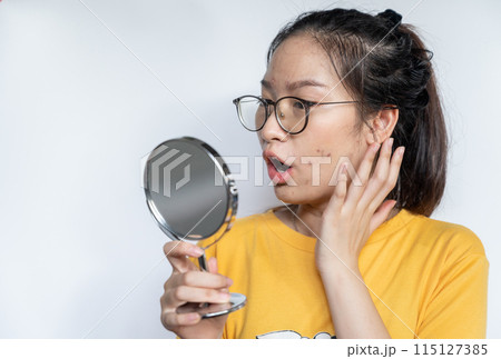 Young Asian woman feeling shock while looking acne and scar occur on face by mini mirror. Conceptual shot of Acne and Problem Skin on female face. 115127385