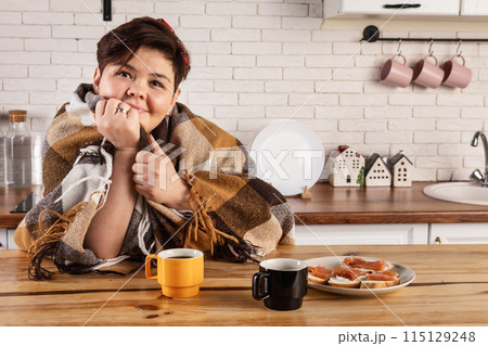 Overweight woman wrapped in a blanket enjoying a cup of coffee in the kitchen Overweight woman wrapped in a blanket enjoying a cup of coffee in the kitchen 115129248