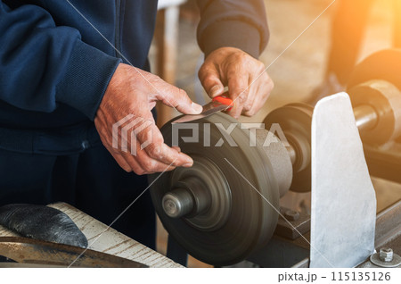 male sharpener grinder sharpens a knife blade on sharpening machine with rotating abrasive wheel close-up 115135126
