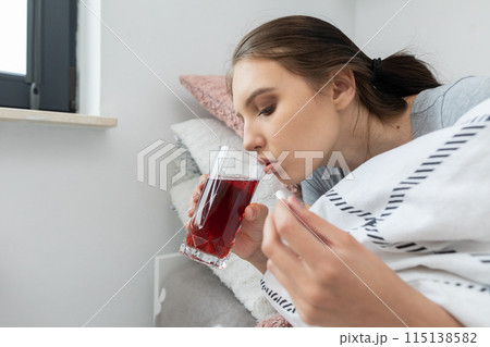 A sick teenager is drinking hot tea with herbs while lying in her bed. 115138582