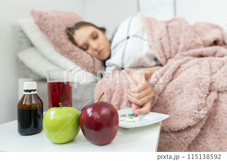 On the bed lies a teenager and in front of her stands a table with apples and medicines that must be swallowed. 115138592