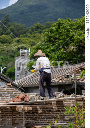 Worker fixing roof in village, wearing straw hat, surrounded by nature and traditional buildings 115142009