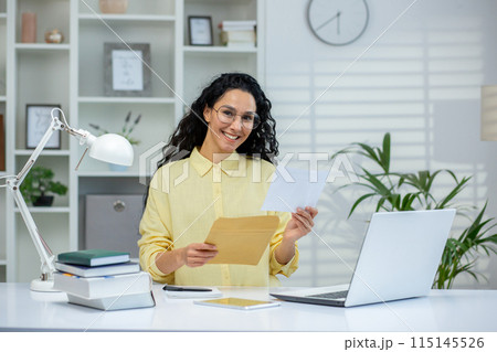 Smiling woman holding an envelope and letter while standing in a bright home office. Happy moment as she reads good news or a positive notification. 115145526