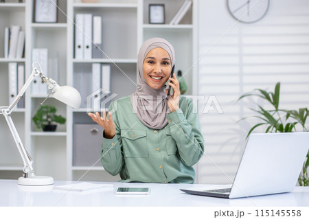 Confident businesswoman wearing a hijab talking on phone, sitting at desk with laptop in bright, modern office environment. Confident businesswoman wearing a hijab talking on phone, sitting at desk with laptop in bright, modern office environment. 115145558