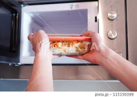 耐熱ガラスのタッパーウェアを電子レンジに入れて温める男性	 115150399