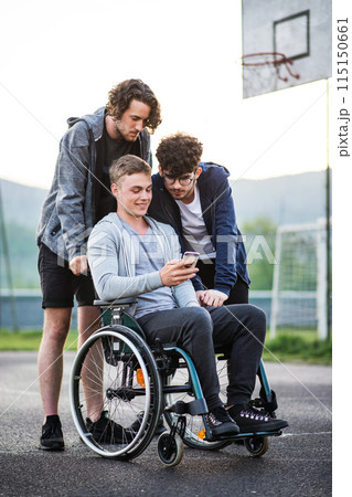 Young man in wheelchair spending free time outdoor with friends, looking at something on smartphone. Male friendship. Young man in wheelchair spending free time outdoor with friends, looking at something on smartphone. Male friendship. 115150661
