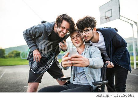 Young man in wheelchair spending free time outdoor with friends, taking selfie, having fun in city. Male friendship. 115150663