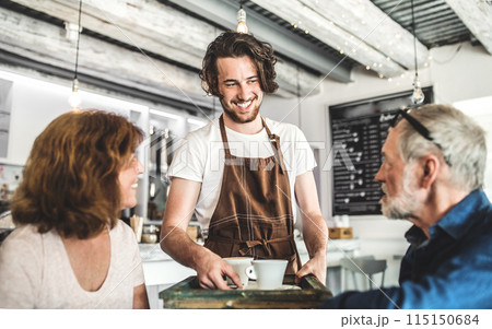 Male barista working in coffee shop, serving customers, elderly couple. University student working part-time in cafe. 115150684