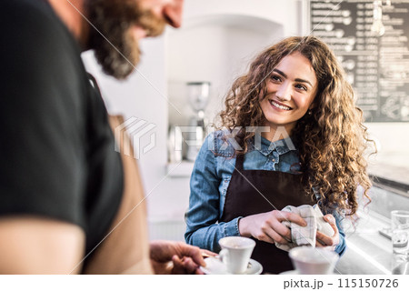 Two young baristas working in coffee shop, standing by counter. Teamwork. 115150726