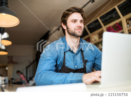 Man in small business, working in his bistro, writing email to supplier. Young male business owner. Man in small business, working in his bistro, writing email to supplier. Young male business owner. 115150758