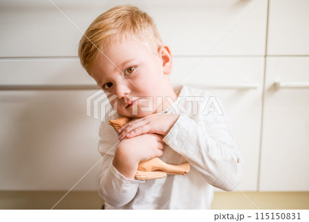 Portrait of a cute boy sitting on the floor in the kitchen, holding wooden horse toy. 115150831