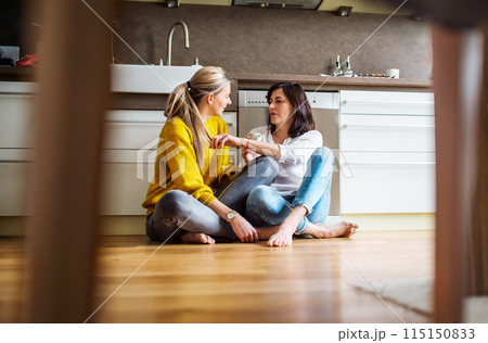 Mother and mature daughter in kitchen, sitting on floor and talking. Closeness of mom and her adult offspring. 115150833