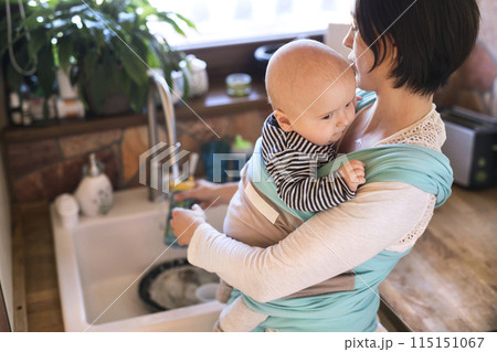 Mother holding small baby while cleaning dishes, carrying him around house in baby wrap. Household chores. Mother holding small baby while cleaning dishes, carrying him around house in baby wrap. Household chores. 115151067