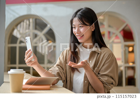 A cheerful Asian woman is talking on a video call or recording her video with her phone in a cafe. 115157023